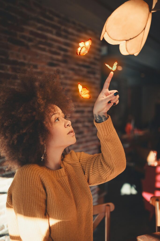 A woman with afro hair in a cozy setting playfully interacts with butterflies indoors.